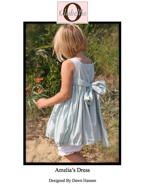 A child wearing a sleeveless light blue dress with a large bow at the back, shown outdoors in a grassy setting. The image includes a pink brand label at the top reading 'Obliterate' and text below reading 'Amelia's Dress' and 'Designed by Dawn Hansen'.
Photo by Olabelhe [image_link]