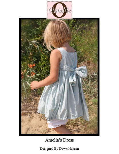 A child wearing a sleeveless light blue dress with a large bow at the back, shown outdoors in a grassy setting. The image includes a pink brand label at the top reading 'Obliterate' and text below reading 'Amelia's Dress' and 'Designed by Dawn Hansen'.
Photo by Olabelhe [image_link]
