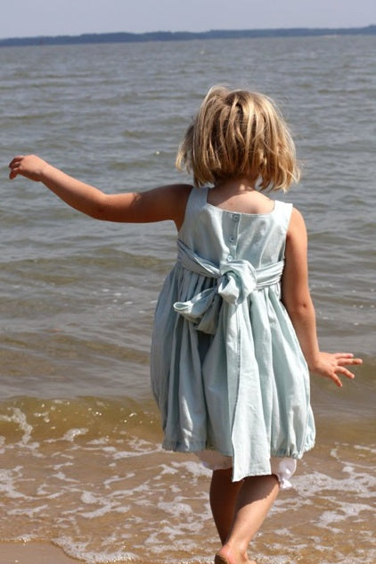 A young girl with blonde hair seen from behind, wearing a light blue sleeveless dress with a tied waist sash, standing on a sandy beach with the ocean water in the background.
Photo by Olabelhe [additional_image_link]