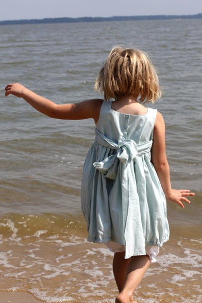 A young girl with blonde hair seen from behind, wearing a light blue sleeveless dress with a tied waist sash, standing on a sandy beach with the ocean water in the background.
Photo by Olabelhe [additional_image_link]