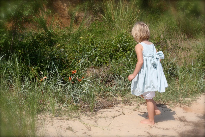 A young child with short blonde hair walks barefoot along a sandy path, viewed from behind, wearing a light blue sleeveless dress with a bow at the back and white shorts, set against a background of green beach grass.
Photo by Olabelhe [additional_image_link]