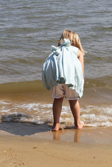 A young child stands barefoot at the edge of the ocean, seen from behind, wearing a light blue sleeveless cover-up or poncho over a pale pink outfit.
Photo by Olabelhe [additional_image_link]