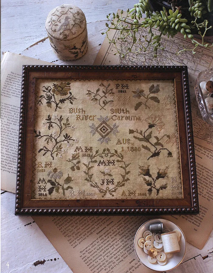 A framed vintage-style cross-stitch sampler with floral embroidery and stitched text, displayed on a rustic tabletop with old books, buttons, thread, and greenery.
Photo by Blackbird Designs [additional_image_link]