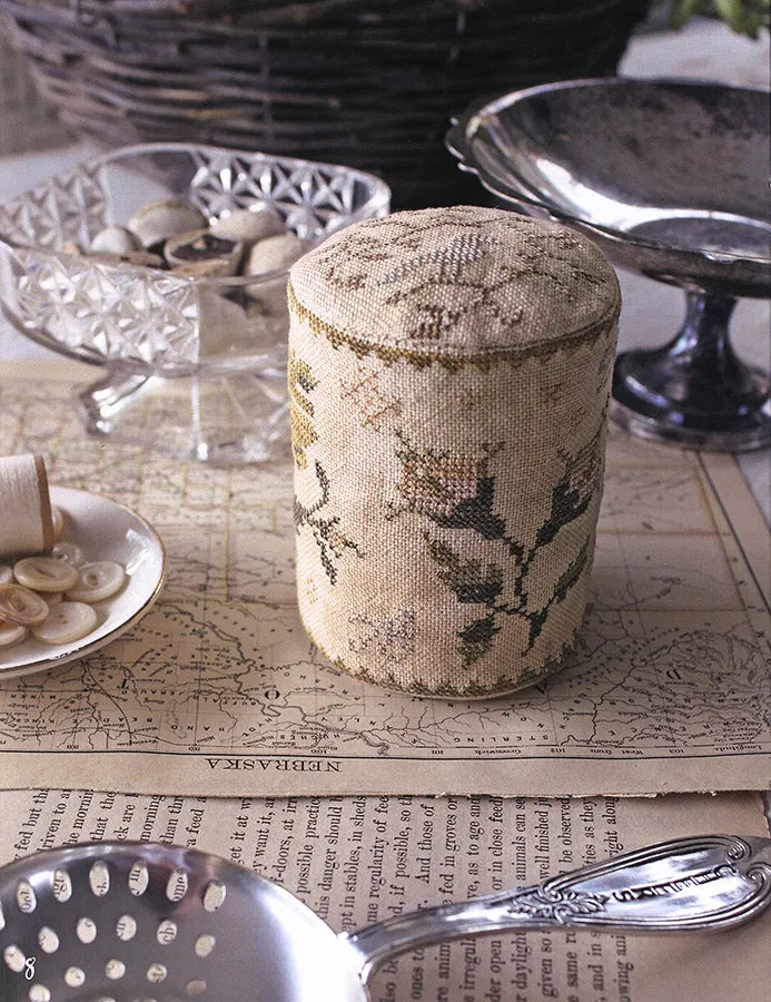 A cylindrical fabric-covered canister or pincushion with a floral embroidered pattern in beige, olive green, and brown, styled on a vintage map surrounded by antique kitchen and household props.
Photo by Blackbird Designs [additional_image_link]