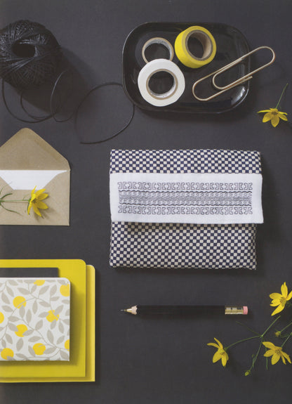A styled flat-lay featuring a black and white checkered fabric pouch with a white embroidered band, surrounded by yellow and black stationery props on a dark gray background.
Photo by Search Press [additional_image_link]