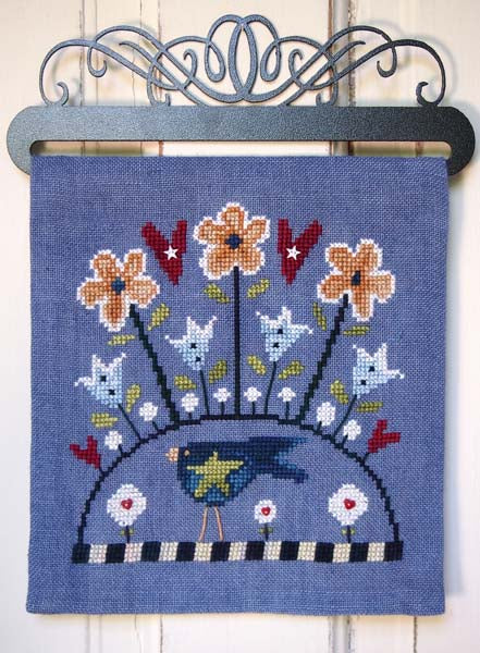 A square blue needlepoint canvas featuring a whimsical design of flowers, birds, and a sun, hung on a black metal hanger against a white wood-paneled wall.
Photo by SamSarah Studio Designs [image_link]