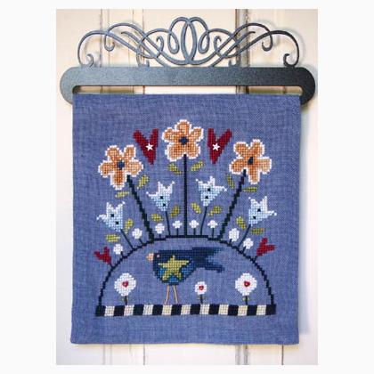 A square blue needlepoint canvas featuring a whimsical design of flowers, birds, and a sun, hung on a black metal hanger against a white wood-paneled wall.
Photo by SamSarah Studio Designs [additional_image_link]