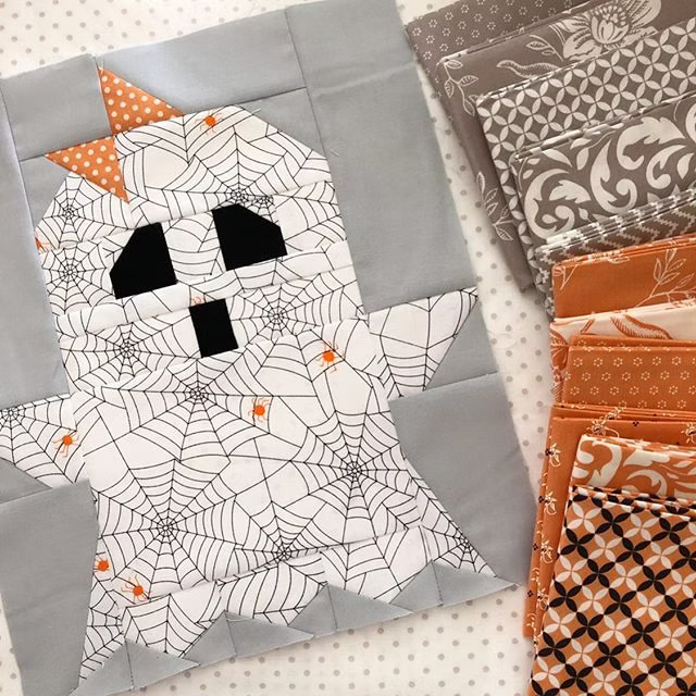 A Halloween-themed quilted fabric piece featuring a white ghost with black spiderweb patterns, black eyes and mouth, and orange accents, displayed next to other patterned fabric swatches.
Photo by ThePatternBasket.com [additional_image_link]