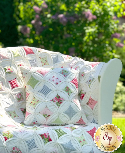 A quilt and pillow set featuring a cathedral window pattern with multicolored patches, showcased on a white chair outdoors.
Photo by Shabby Fabrics [additional_image_link]