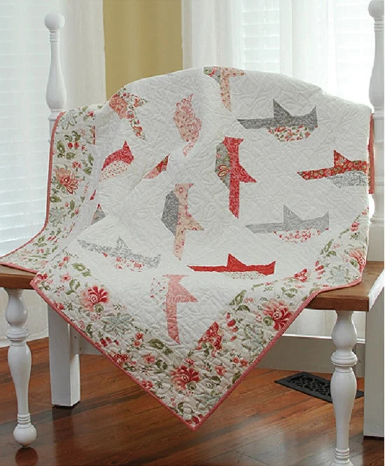 A quilted baby blanket draped over a white wooden chair in a bright room. The blanket features a white background with red and gray paper airplane motifs and a red floral border.
Photo by ThePatternBasket.com [additional_image_link]