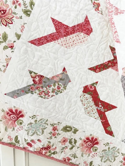 A close-up of a quilted textile featuring stylized bird shapes in red, pink, gray, and floral print fabrics, with a white quilted center and a floral border.
Photo by ThePatternBasket.com [additional_image_link]