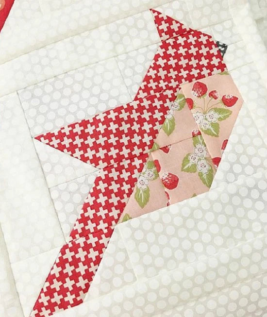 A close-up of a quilted fabric block featuring a bird motif made from three patterned fabrics: red and white crosshatch, red and white strawberry print, and light pink with strawberry and leaf floral print, all set on a white polka-dotted quilt background.
Photo by ThePatternBasket.com [additional_image_link]