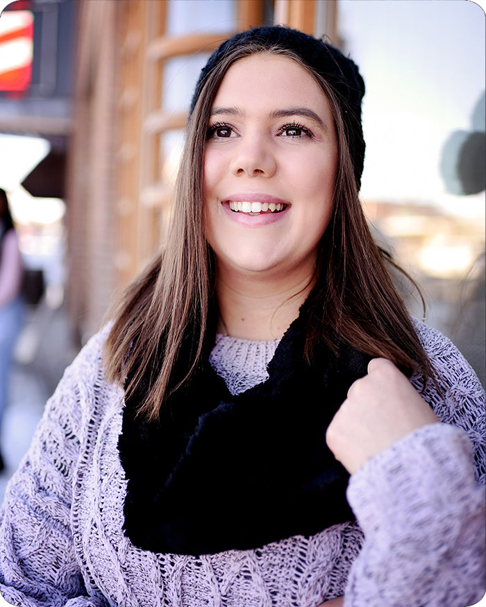 A smiling young woman wearing a black knit beanie, a black faux-fur neck warmer, and a lavender-gray cable-knit sweater, photographed outdoors with a blurred background.
Photo by Shannon Fabrics [additional_image_link]