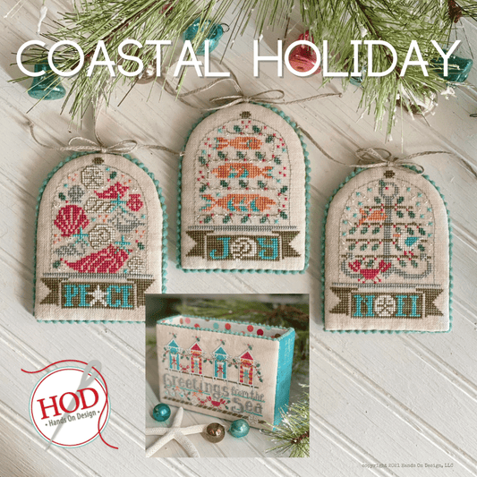 Three cross stitch embroidery ornaments with coastal and beach themes, and a storage box with the text 'Coastal Holiday' on a white background. The ornaments are hung by small green pine branches.
Photo by Handsondesign.com [image_link]