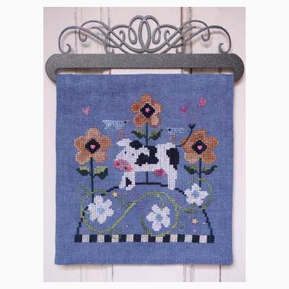 Decorative cross stitch with a cow and flowers on a blue background, hanging on a metal rack.
Photo by SamSarah Studio Designs [image_link]