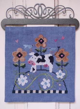 Decorative cross stitch with a cow and flowers on a blue background, hanging on a metal rack.
Photo by SamSarah Studio Designs [additional_image_link]