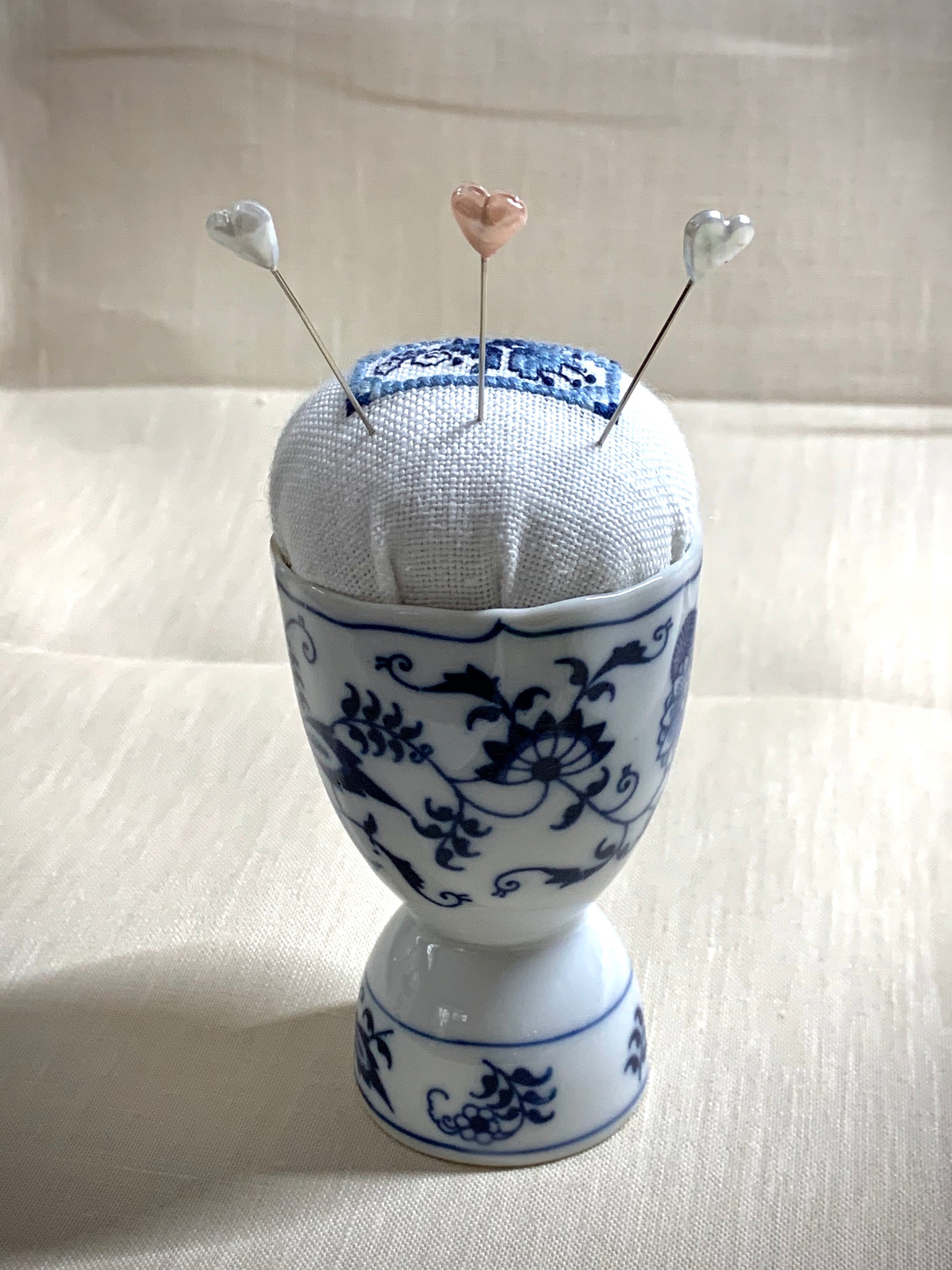 A close-up of a hand-stitched Delft pincushion with a vintage blue and white Danube double egg cup motif on white linen.
Photos by The Surgeon's Knots [additional_image_link]