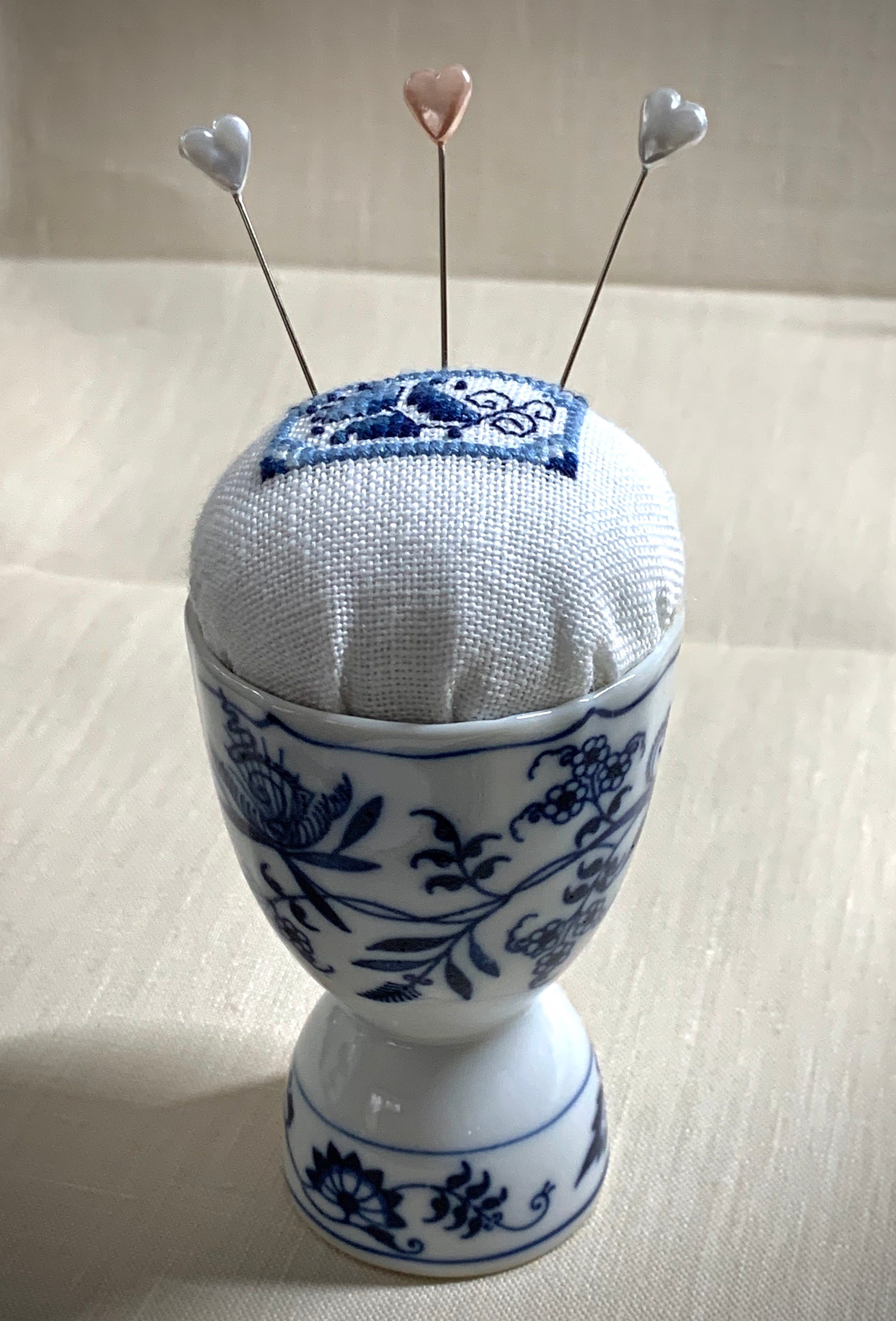 A close-up of a hand-stitched Delft pincushion with a vintage blue and white Danube double egg cup motif on white linen.
Photos by The Surgeon's Knots [additional_image_link]