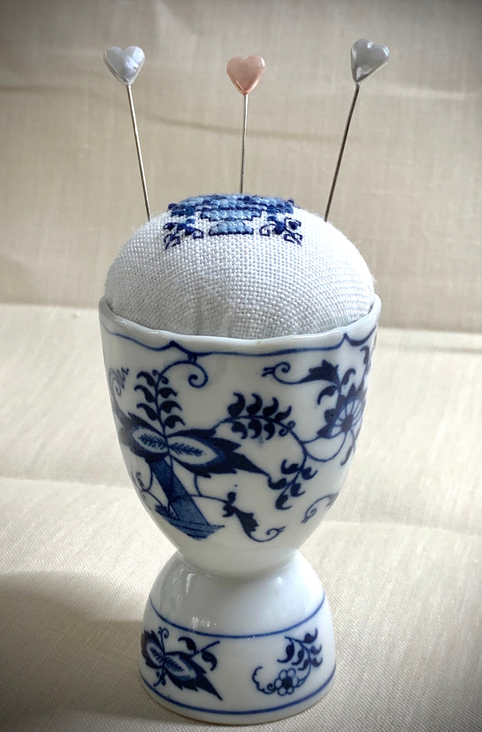 A close-up of a hand-stitched Delft pincushion with a vintage blue motif on white linen, featuring a blue and white porcelain double egg cup design.
Photos by The Surgeon's Knots [additional_image_link]