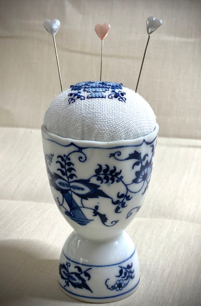 A close-up of a hand-stitched Delft pincushion with a vintage blue motif on white linen, featuring a blue and white porcelain double egg cup design.
Photos by The Surgeon's Knots [additional_image_link]