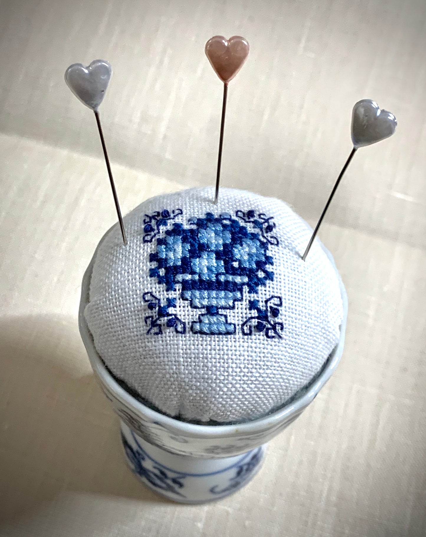 A close-up of a hand-stitched Delft pincushion with a vintage blue motif on white linen, featuring a blue and white porcelain double egg cup design.
Photos by The Surgeon's Knots [additional_image_link]