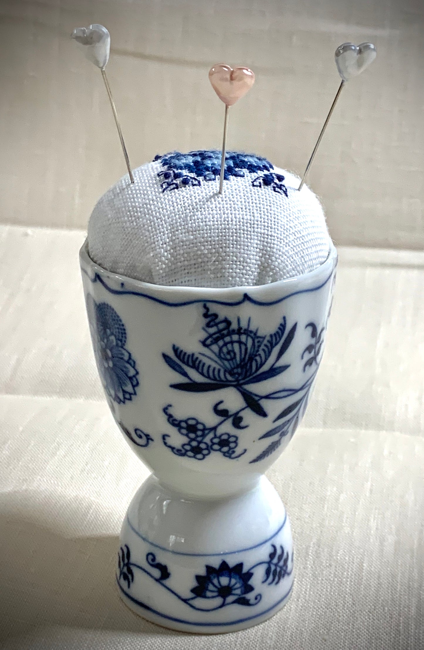 A close-up of a hand-stitched Delft pincushion with a vintage blue motif on white linen, featuring a blue and white porcelain double egg cup design.
Photos by The Surgeon's Knots [additional_image_link]