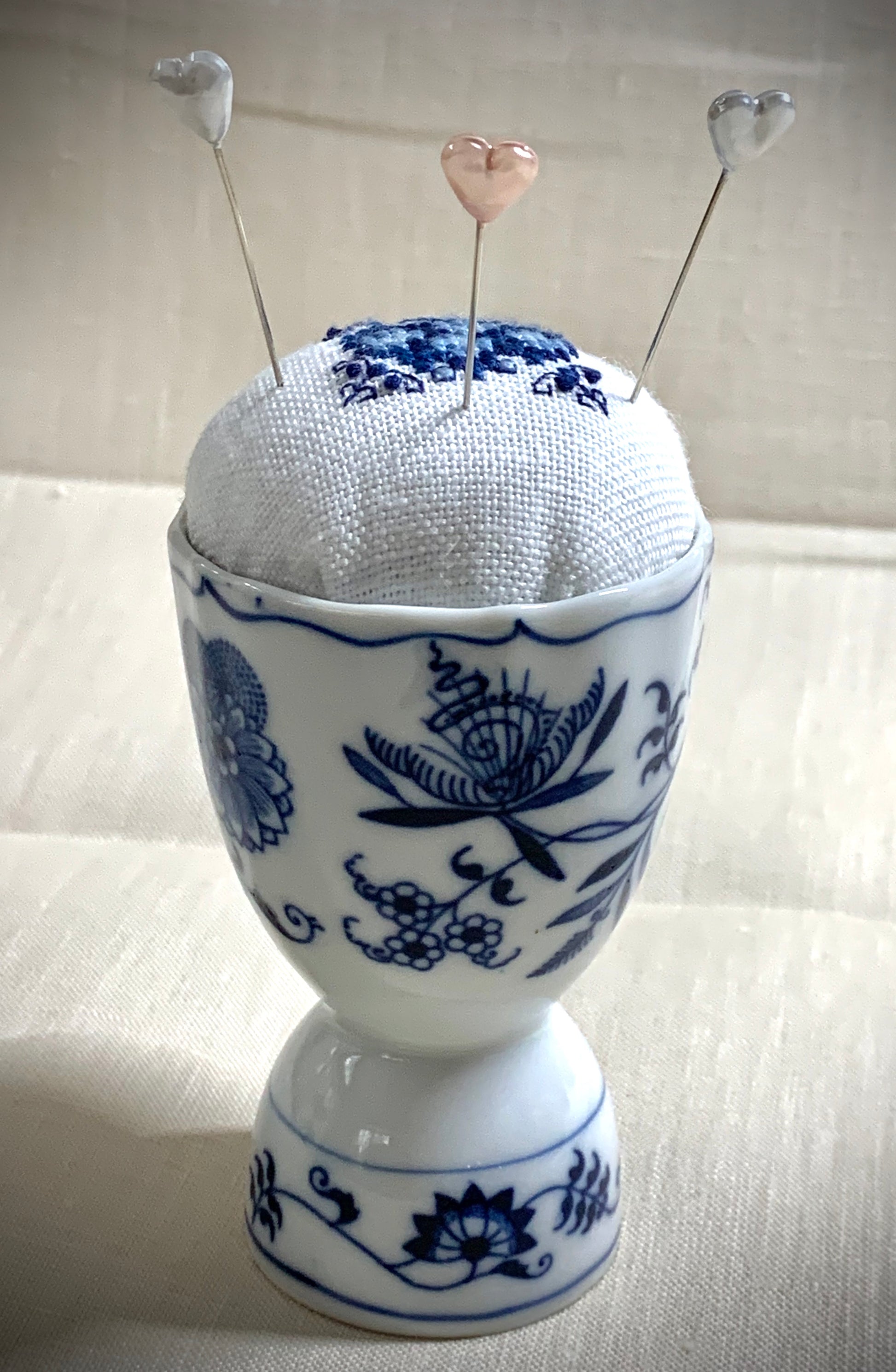 A close-up of a hand-stitched Delft pincushion with a vintage blue motif on white linen, featuring a blue and white porcelain double egg cup design.
Photos by The Surgeon's Knots [additional_image_link]