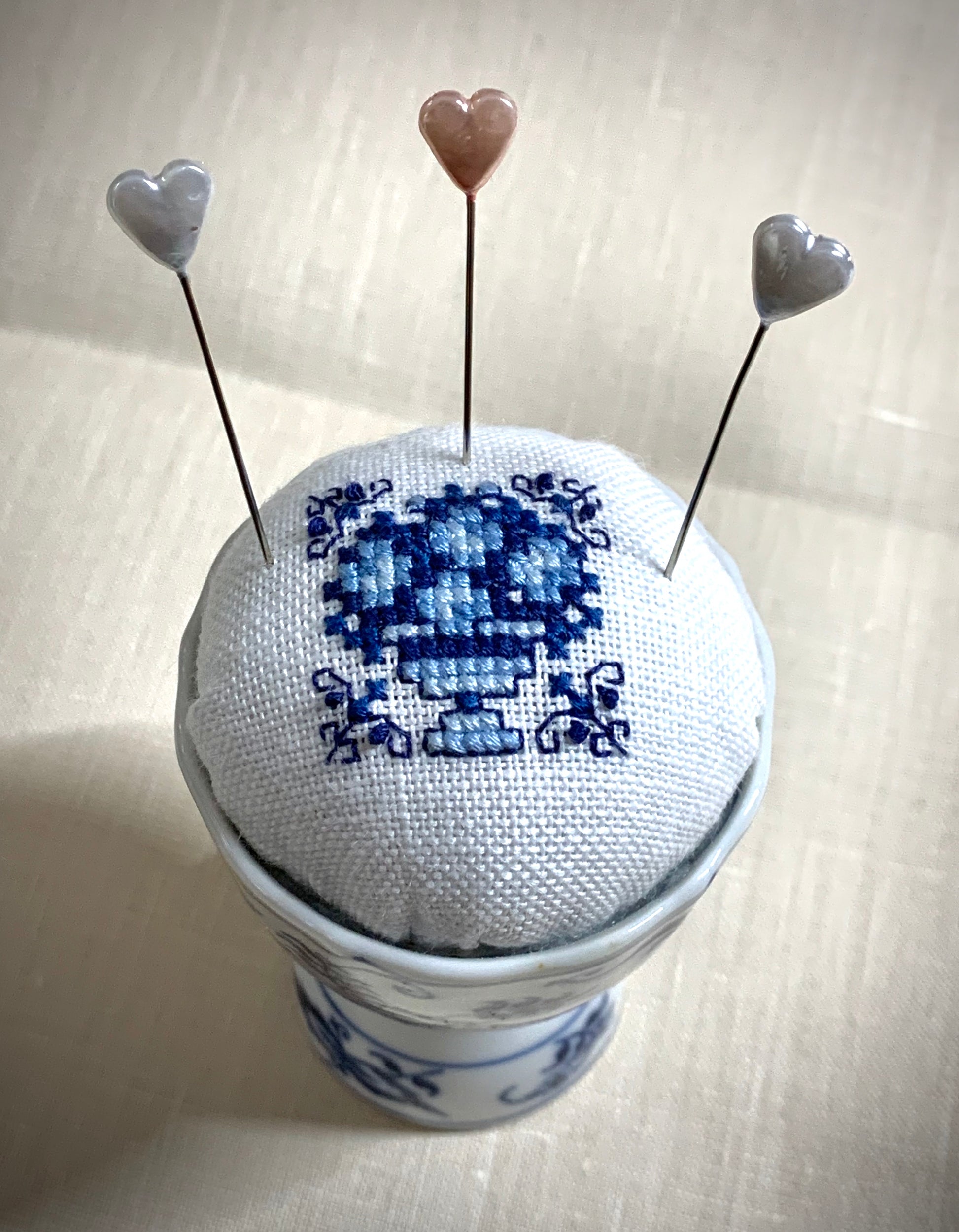 A close-up of a hand-stitched Delft pincushion with a vintage blue motif on white linen, featuring a blue and white porcelain double egg cup design.
Photos by The Surgeon's Knots [additional_image_link]