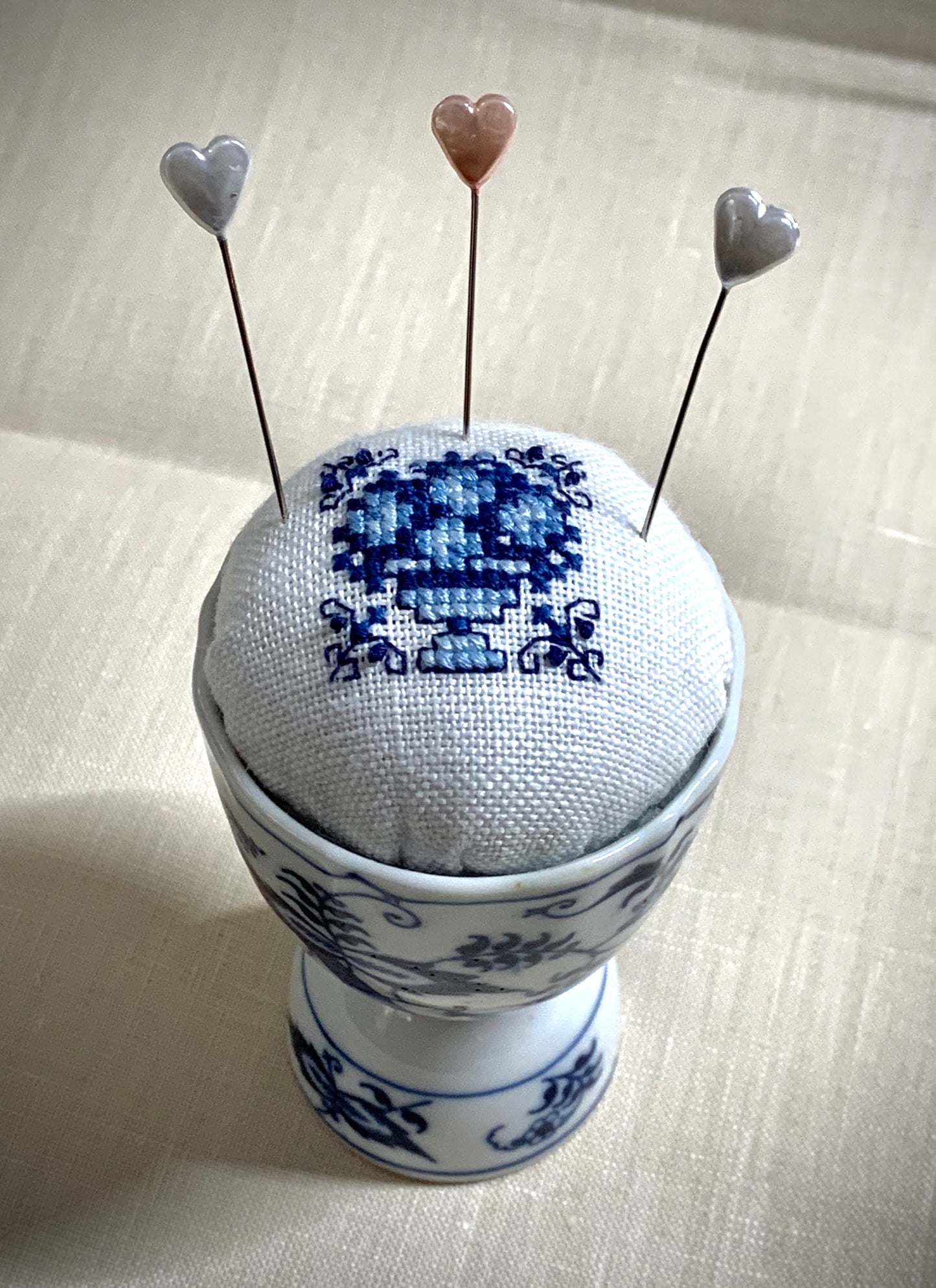 A close-up of a hand-stitched Delft pincushion with a vintage blue motif on white linen, featuring a blue and white porcelain double egg cup design.
Photos by The Surgeon's Knots [additional_image_link]