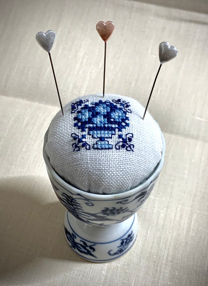 A close-up of a hand-stitched Delft pincushion with a vintage blue motif on white linen, featuring a blue and white porcelain double egg cup design.
Photos by The Surgeon's Knots [additional_image_link]