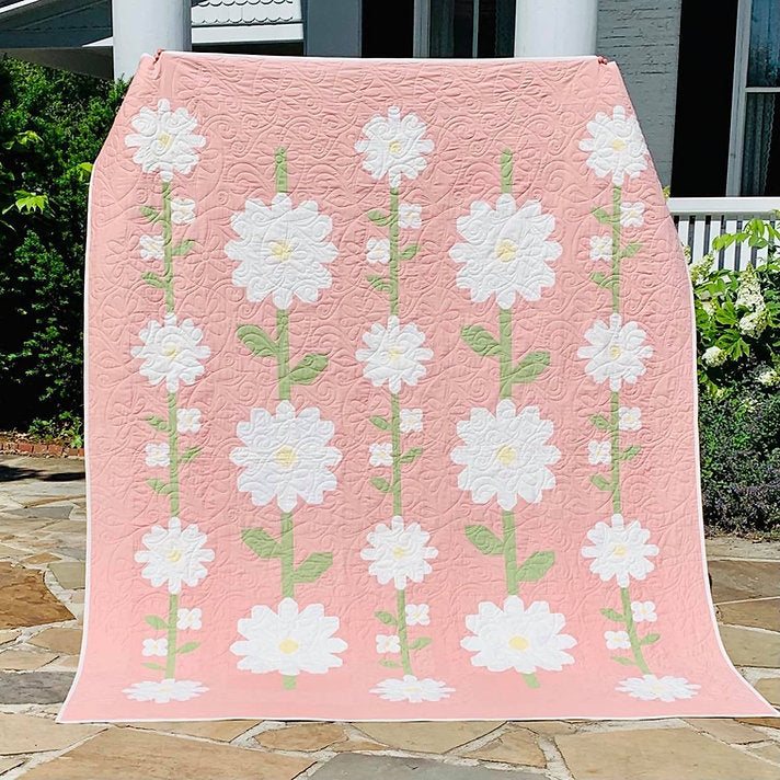 A pink quilted blanket or throw featuring a repeating pattern of large white daisy-like flowers with green stems, draped outdoors over a patio railing with plants and a house visible in the background.
Photo by My Sew Quilty Life [additional_image_link]