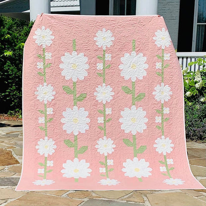 A pink quilted blanket or throw featuring a repeating pattern of large white daisy-like flowers with green stems, draped outdoors over a patio railing with plants and a house visible in the background.
Photo by My Sew Quilty Life [additional_image_link]