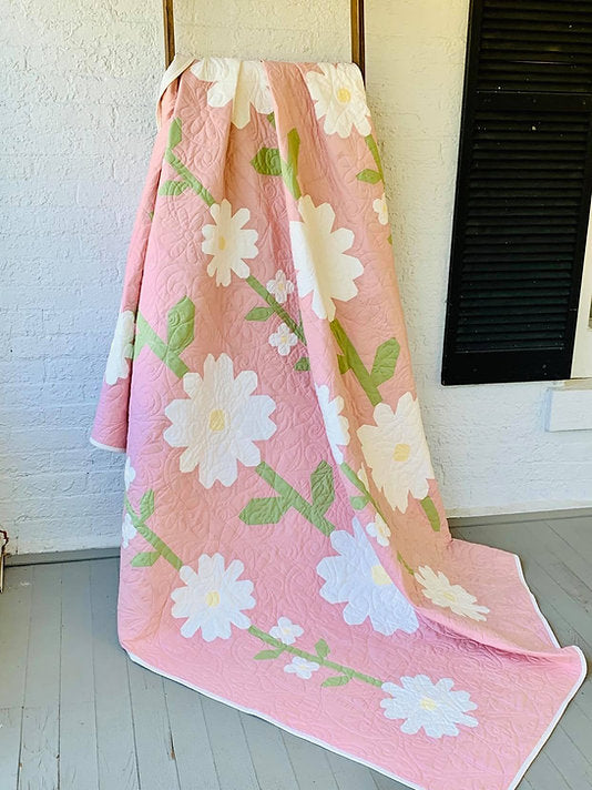A pink quilted blanket with a white daisy floral pattern draped over a chair in a bright room with a white brick wall and light wood floor.
Photo by My Sew Quilty Life [additional_image_link]