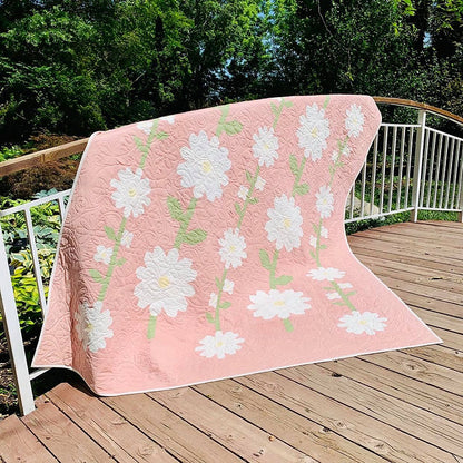 A pink quilted blanket with a white daisy floral pattern draped over a white metal railing on a wooden deck, with green trees in the background.
Photo by My Sew Quilty Life [additional_image_link]