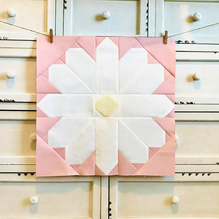 A square quilted fabric block featuring a stylized flower design in shades of pink and white, with a pale yellow center, hanging from two wooden clips against a white wooden drawer chest.
Photo by My Sew Quilty Life [additional_image_link]