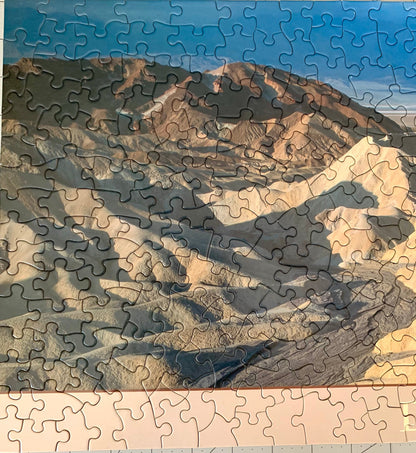 A jigsaw puzzle depicting a panoramic view of the Death Valley landscape, including the Zabriskie Point area, with a blue sky and distant mountains.
Photo by Karen King [additional_image_link]