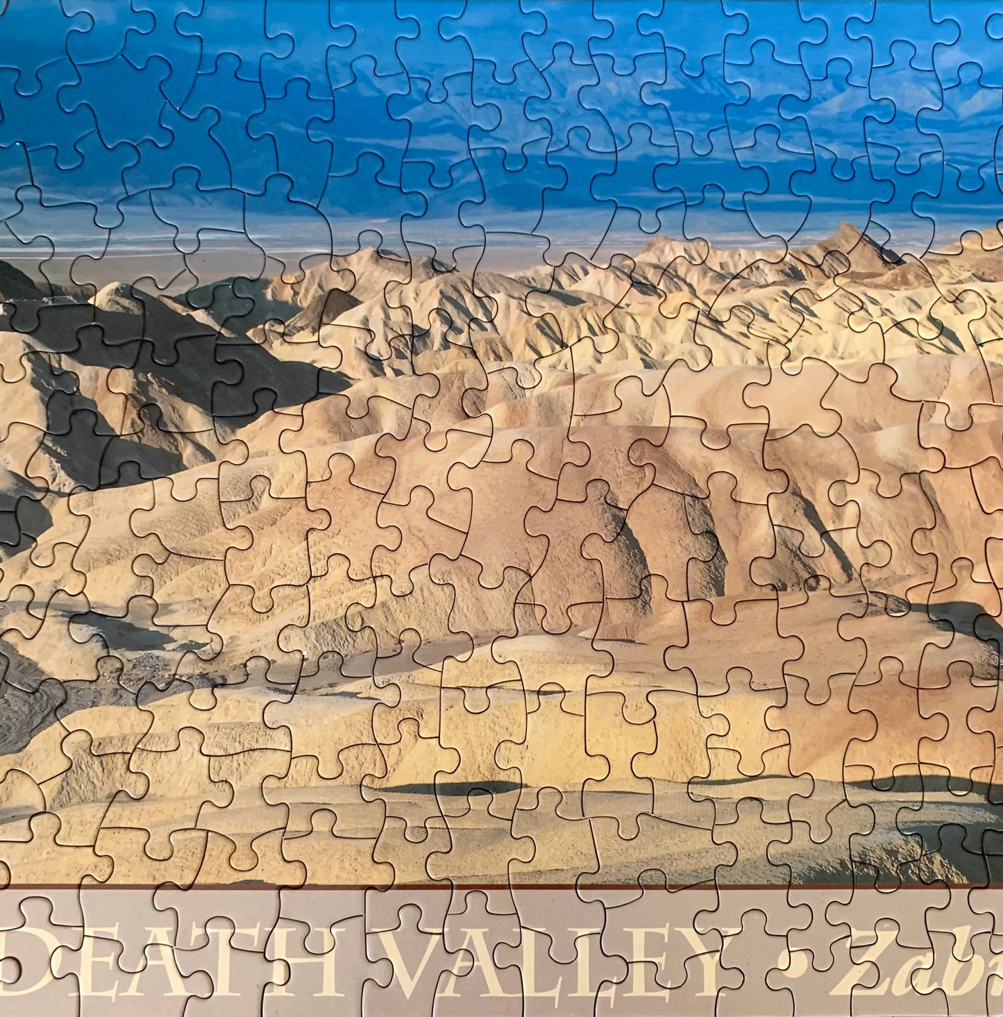 A jigsaw puzzle depicting a panoramic view of the Death Valley landscape, including the Zabriskie Point area, with a blue sky and distant mountains.
Photo by Karen King [additional_image_link]