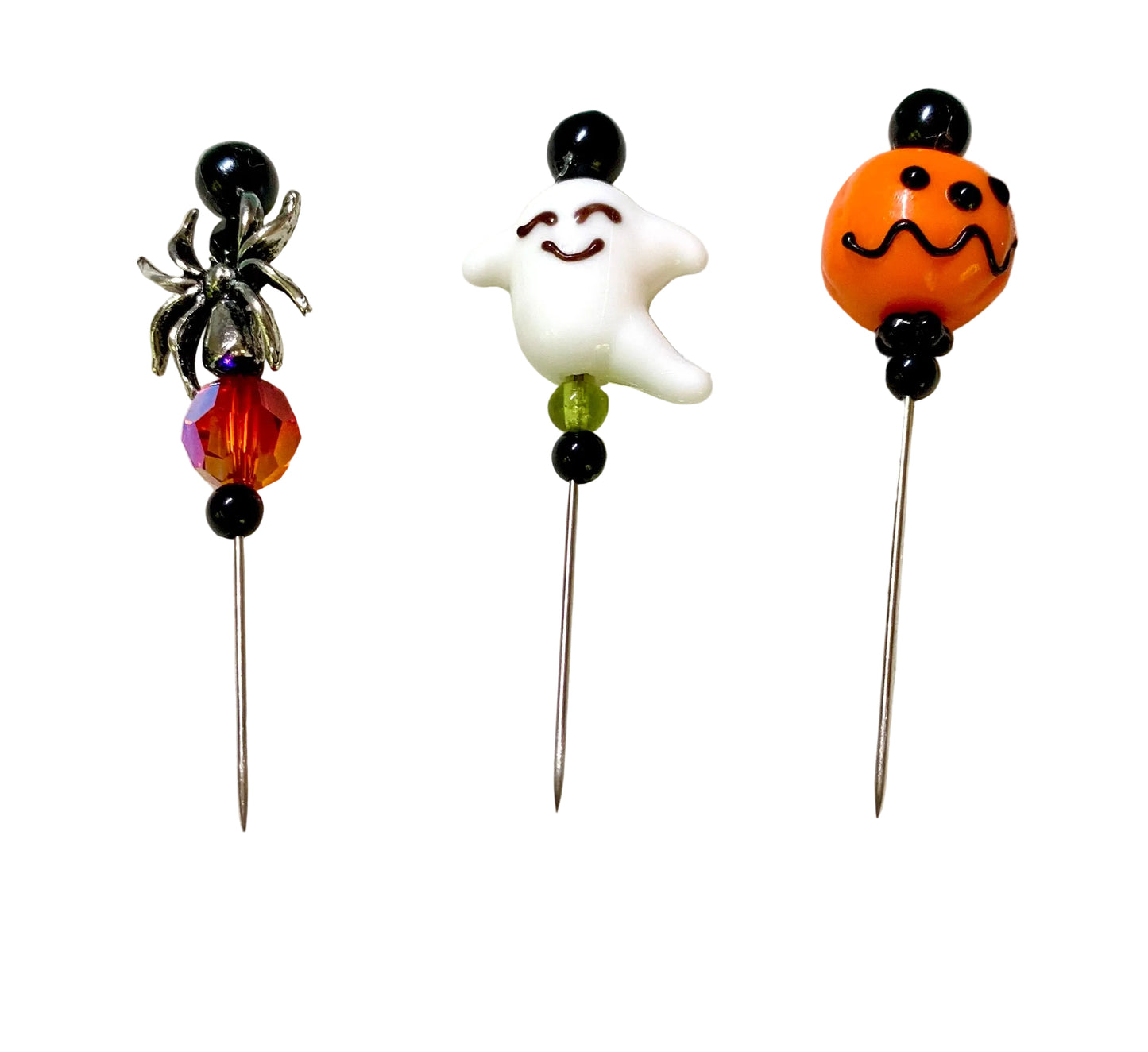 Three Halloween-themed glass beads on metal pins, featuring a black spider, a smiling white ghost, and an orange jack-o'-lantern head with black beads and faceted glass accents, set against a plain white background.
[additional_image_link]