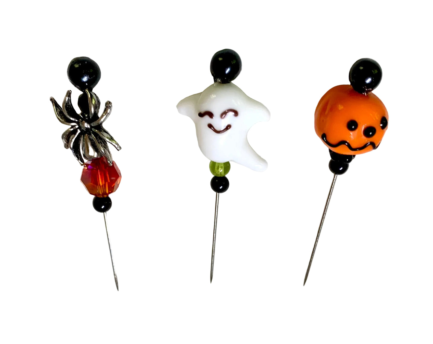 Three Halloween-themed glass beads on metal pins, featuring a silver spider with an amber-red bead, a white smiling ghost with a small green bead, and an orange jack-o'-lantern with black facial details.
[additional_image_link]