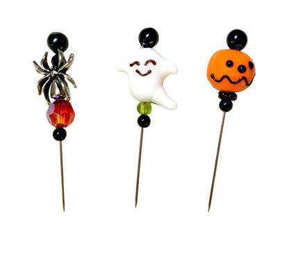 Three Halloween-themed beads on metal pins, featuring a silver spider, a white smiling ghost, and an orange jack-o'-lantern bead, displayed on a white background.
[image_link]