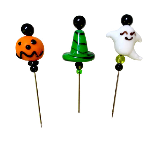 Three Halloween-themed glass pins on metal needles, featuring an orange jack-o'-lantern, a green witch hat with black stripes, and a white smiling ghost, all set against a plain white background.
[image_link]
