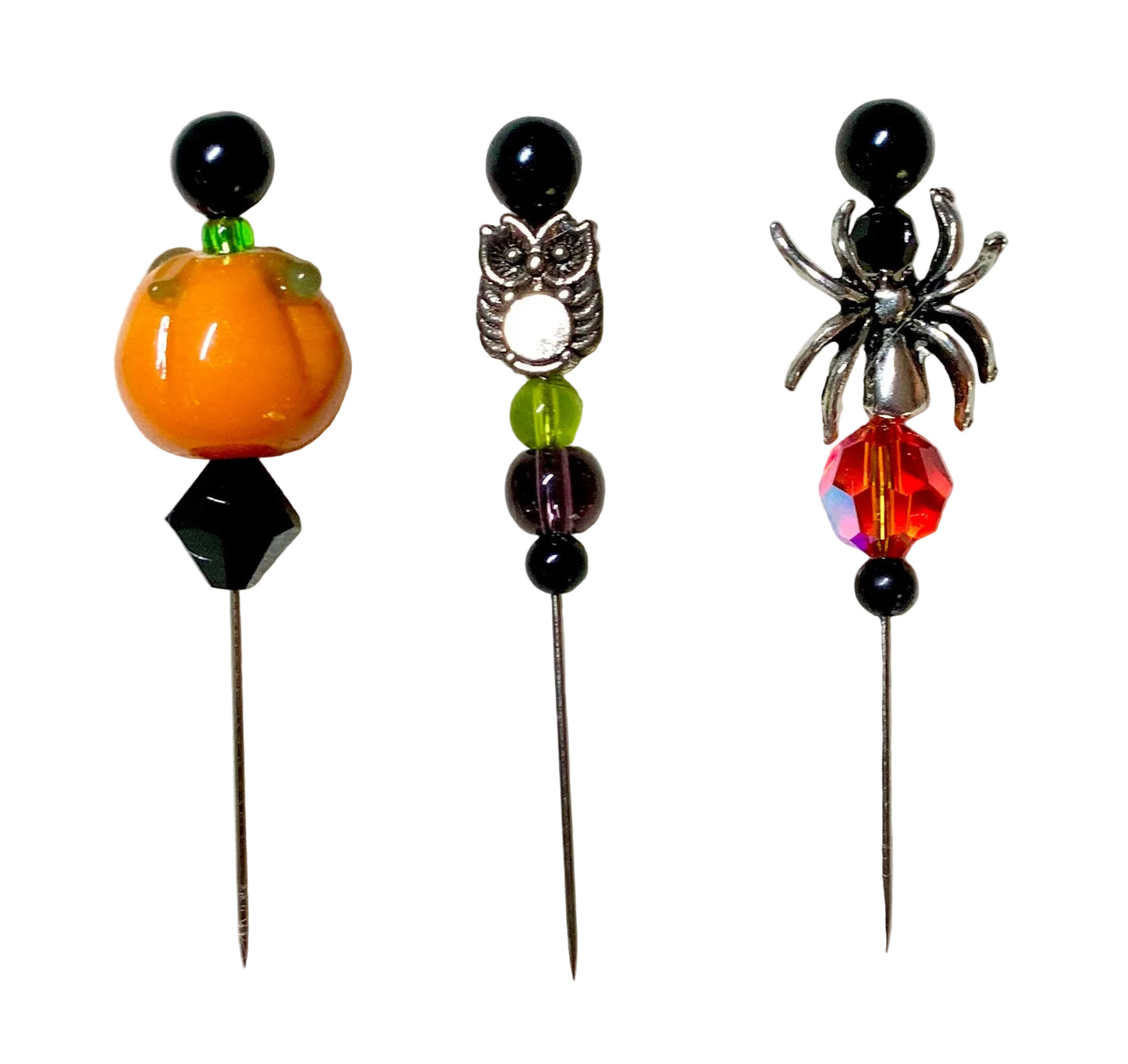 Three Halloween-themed beaded pins on a white background. The left pin features an orange pumpkin with green leaves and a black bead. The right pin has a silver spider with a fire bead. The middle bead has a silver owl with purple and green beads.
[image_link]