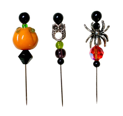Three Halloween-themed beaded pins on a white background. The left pin features an orange pumpkin with green leaves and a black bead. The right pin has a silver spider with a fire bead. The middle bead has a silver owl with purple and green beads.
[image_link]