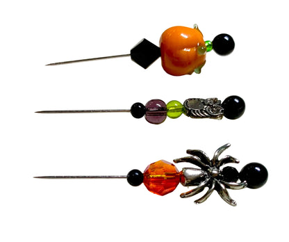 Three Halloween-themed pins with beads and metal charms, displayed on a white surface. The pins feature orange pumpkin, purple and orange beads, a silver owl, and a silver spider charm.
[additional_image_link]