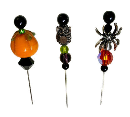 Three Halloween-themed beaded pins on a textured white background. The left pin features an orange pumpkin bead with green stems and black beads. The middle has a silver owl charm and the right has a silver spider charm.
[additional_image_link]