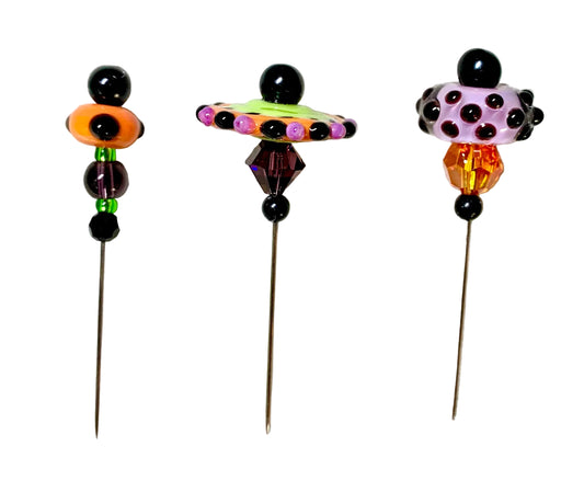 Three decorative beaded pins with long metal needles, displayed on a textured white surface. The pins feature colorful glass beads in orange, purple, black, green, and yellow.
[image_link]