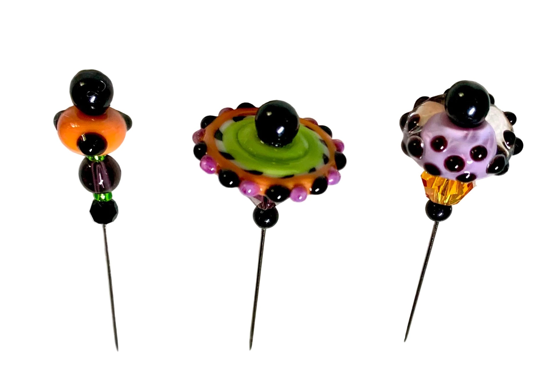 Three handmade beaded pins with long metal needles are arranged on a white surface. Each pin features a unique colorful glass bead design with black beads and small accent beads in purple, green, orange, and yellow.
[additional_image_link]