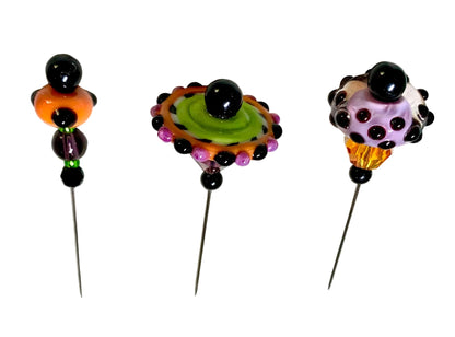 Three handmade glass bead pins with long metal needles, displayed on a white surface. The pins feature colorful, patterned glass beads including purple, green, orange, black, and pink.
[additional_image_link]