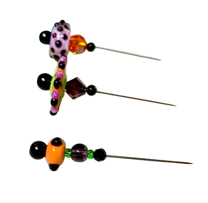 Three handmade beaded pins with metal needles, featuring colorful glass beads in orange, purple, pink, green, and black, arranged on a white mat.
[additional_image_link]