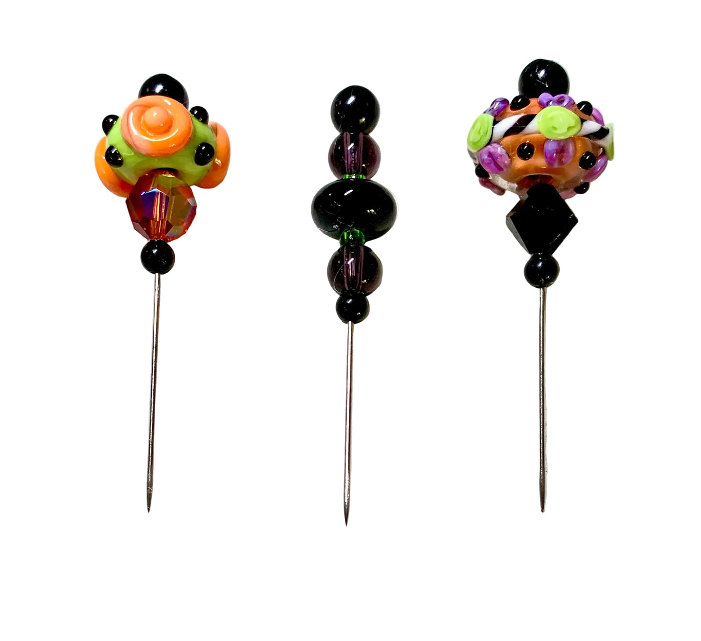 Three handmade glass beads with long metal pins displayed on a white surface. The left pin features a bright orange and green floral bead with black accents, the center pin has a stack of glossy black beads, and the right pin shows a multicolor floral bead with purple, green, and orange details.
[additional_image_link]